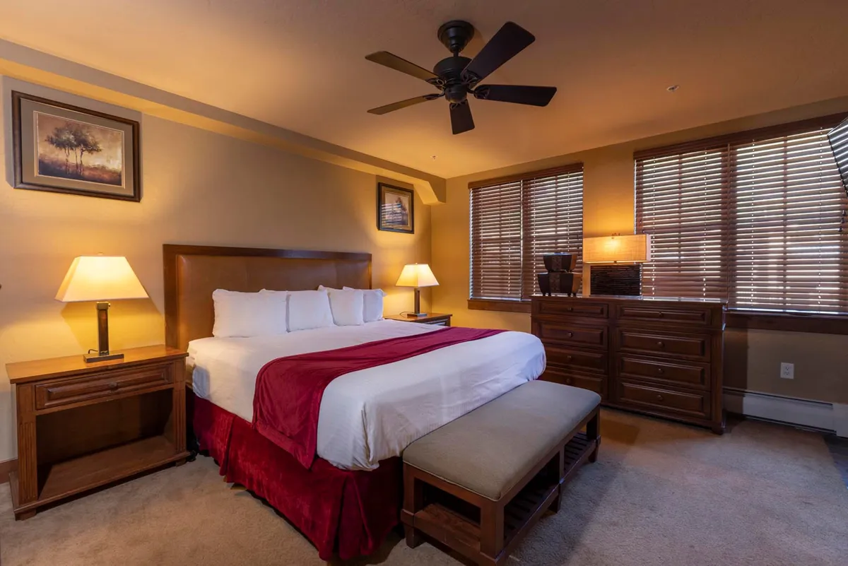 A serene bedroom scene with a well-made bed, stylish nightstands, a dressing table, large windows, and tasteful artwork. Soft morning light floods the room, and the comfortable setting encourages relaxation and enjoying a quiet coffee by the window.