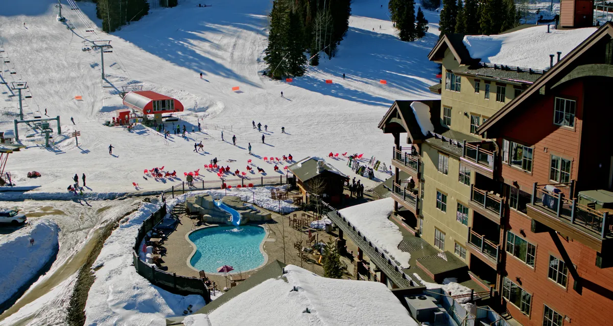 A sweeping exterior view of Purgatory Lodge, with the snow-covered ski slopes and ski lifts just steps from the lodge. The image also features an inviting outdoor heated swimming pool surrounded by the wintry landscape, with guests relaxing both by the pool and at the lodge. The Needles Mountain Range rises in the background, framing the scene with dramatic alpine views.