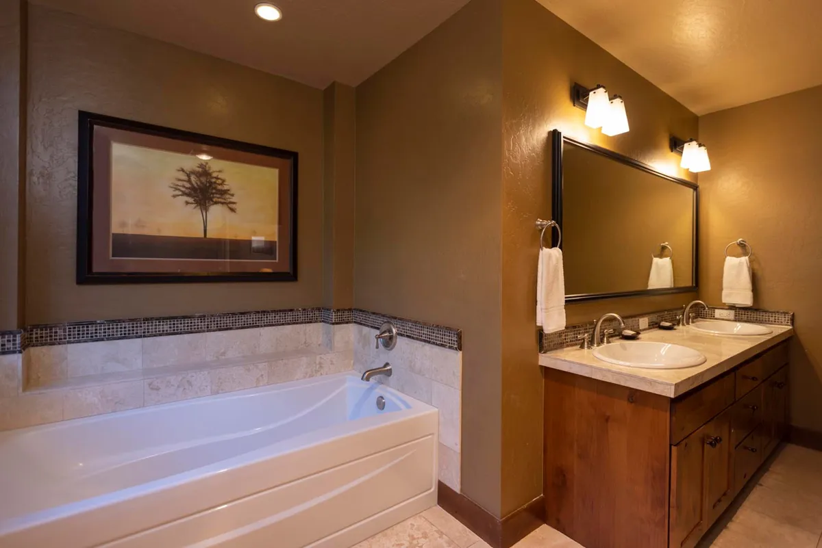 A serene bathroom featuring a bathtub, double sinks, a spacious mirror, and tasteful artwork. The space is designed for relaxation and self-care, evoking a spa-like environment ideal for unwinding.