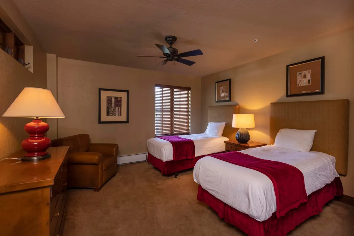 A bright and inviting twin bedroom with two beds, a soft lamp between them, an armchair by the window, and a view of the outdoors. The setup is ideal for siblings, friends, or kids, providing a restful and versatile sleeping space.