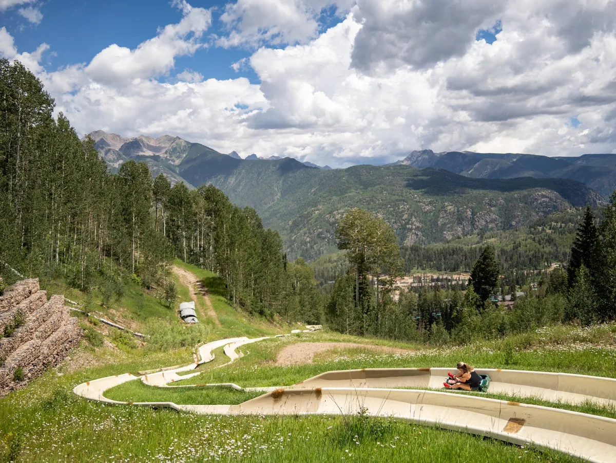 The image captures an exciting slide path set against a stunning mountain backdrop, with lush greenery and a person seated nearby. The scene is lively and playful, suggesting outdoor adventure and family-friendly fun in a scenic setting.