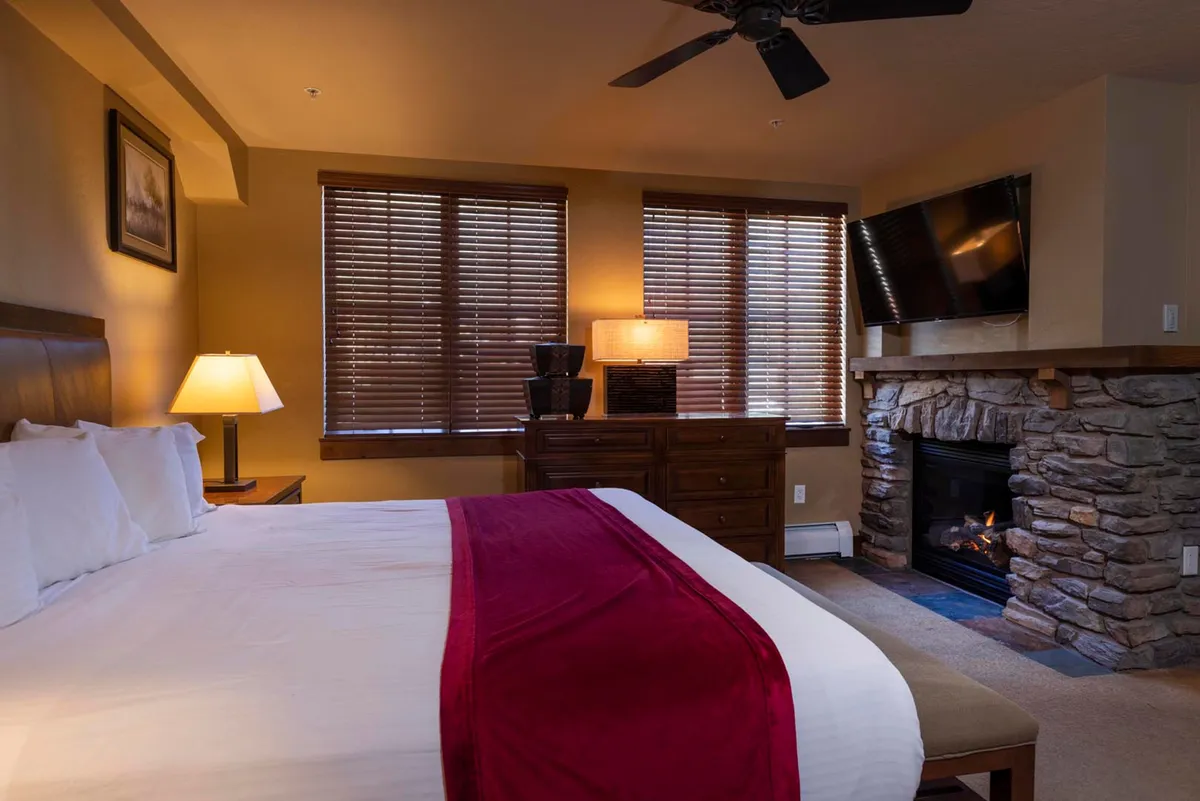 A luxurious bedroom featuring a plush king-size bed, a cozy fireplace at the foot of the bed, large windows bringing in natural light, bedside lamps, and a mounted television. The ambiance is intimate and inviting, perfect for unwinding after a day outdoors.