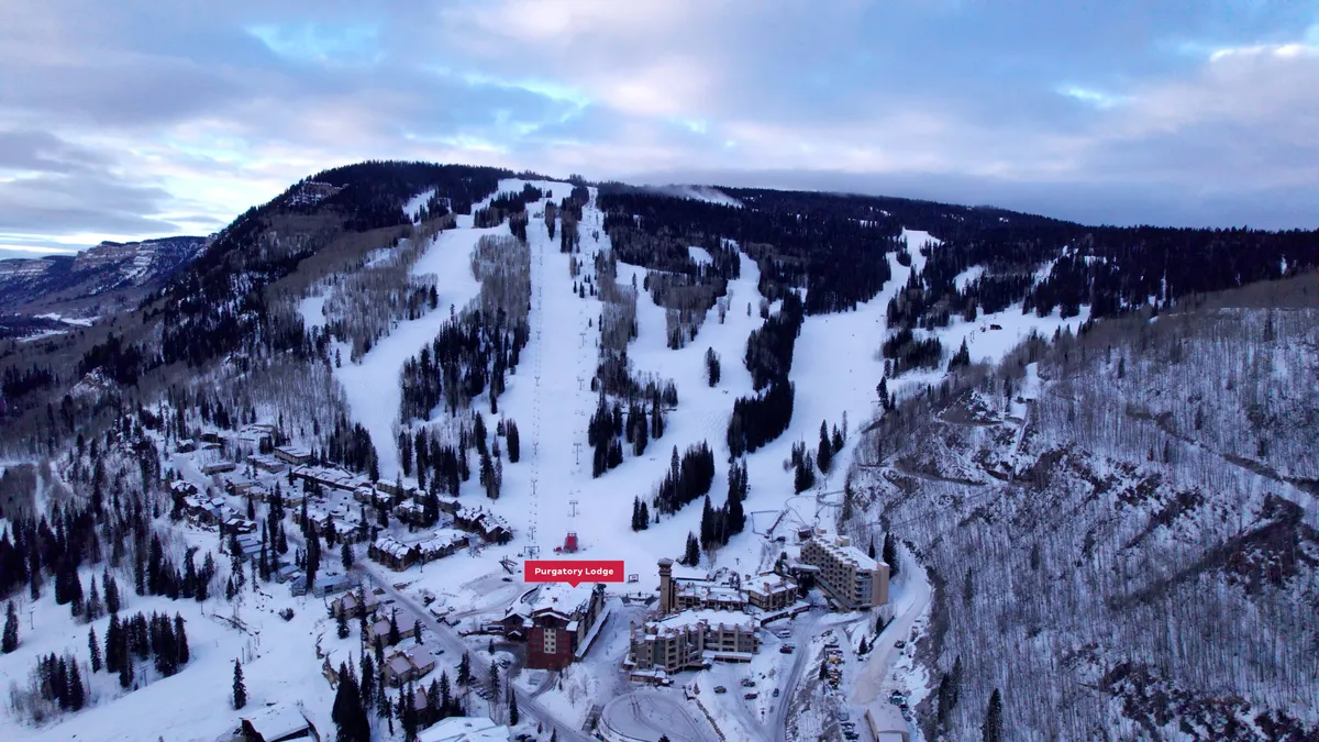 An exterior shot focused on Purgatory Lodge nestled at the base of ski slopes, surrounded by snow-covered mountains and a tree line. The scene includes visible ski trails and a welcoming lodge atmosphere, evoking the excitement of a day on the mountain.