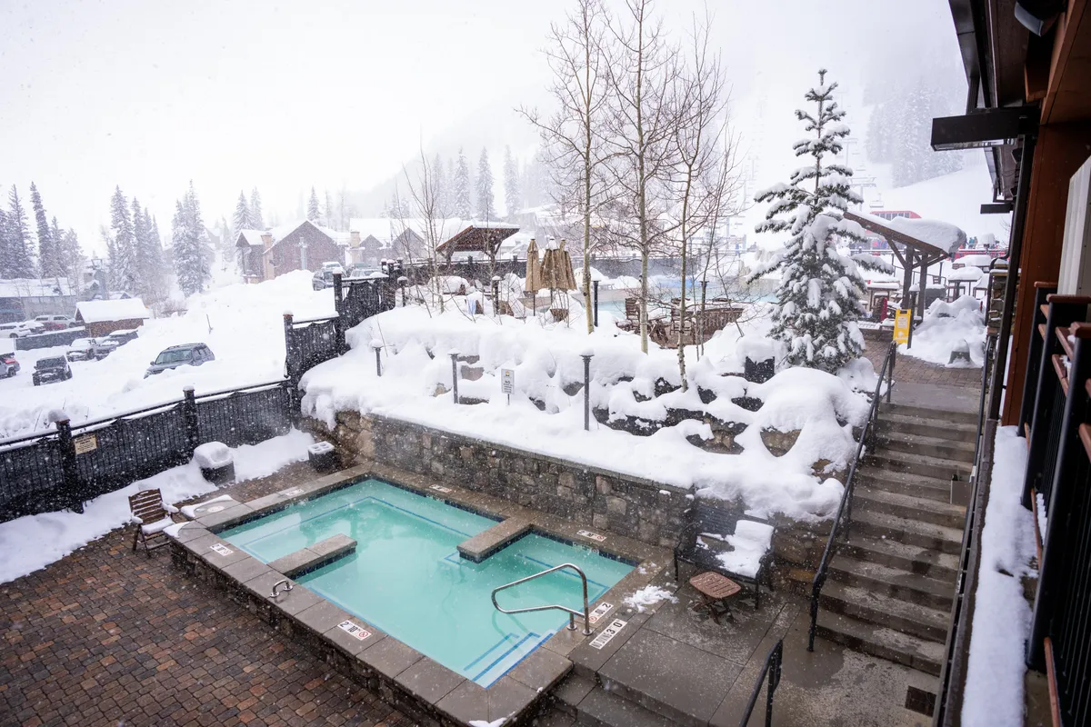 An outdoor hot tub is set against a backdrop of snow-covered trees and landscape. Guests are shown enjoying the warm waters and the scenery, evoking tranquility and indulgence after a day of activity.