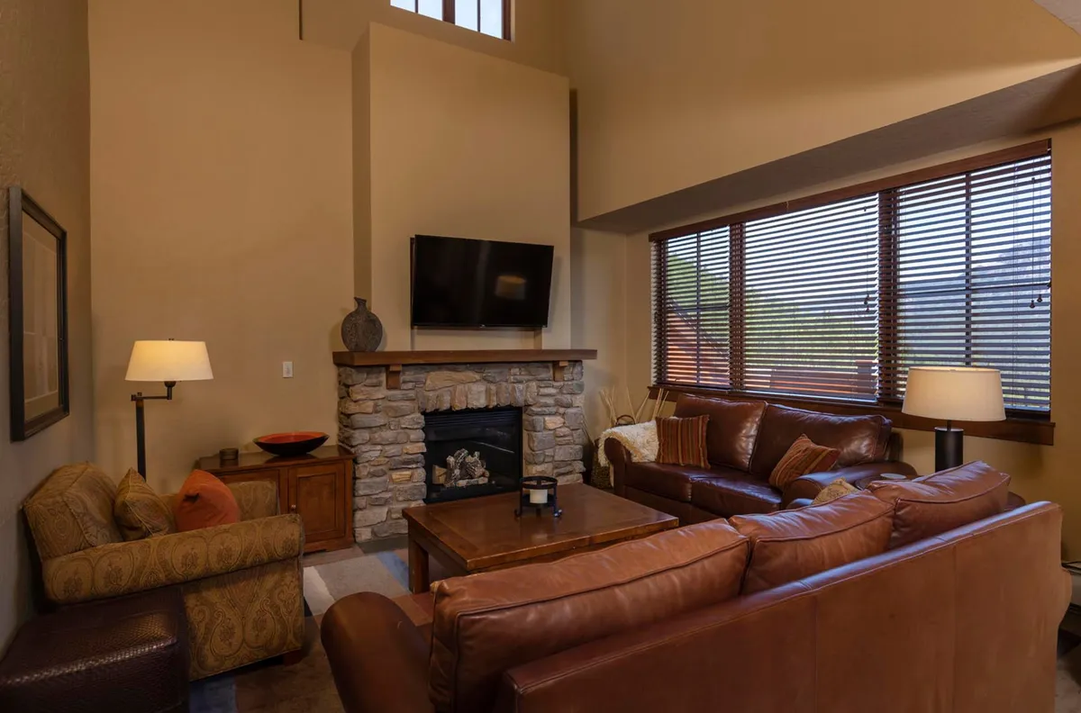 The image showcases a spacious living room with a beautiful stone fireplace as the centerpiece, surrounded by plush leather sofas. Large windows with blinds let in natural light and offer glimpses of the mountain scenery outside, creating a warm and inviting atmosphere perfect for gatherings, relaxation, or simply unwinding after a day on the slopes.