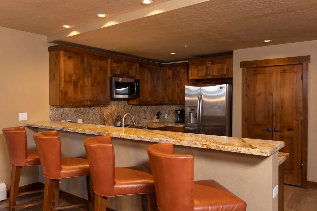 A warm and welcoming kitchen featuring a large granite countertop, wooden cabinetry, bar stools, and gleaming stainless steel appliances. The open layout encourages socializing, with ample space for cooking and gathering. Morning light streams through nearby windows, highlighting the inviting atmosphere ideal for shared meals and celebrations.