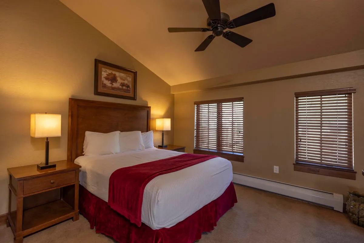 A welcoming bedroom with a bed draped in a red accent blanket, wooden bedside tables, windows with blinds, and framed artwork. The atmosphere is tranquil, with sunlight streaming in to create a cozy and inviting space perfect for morning routines or restful evenings.
