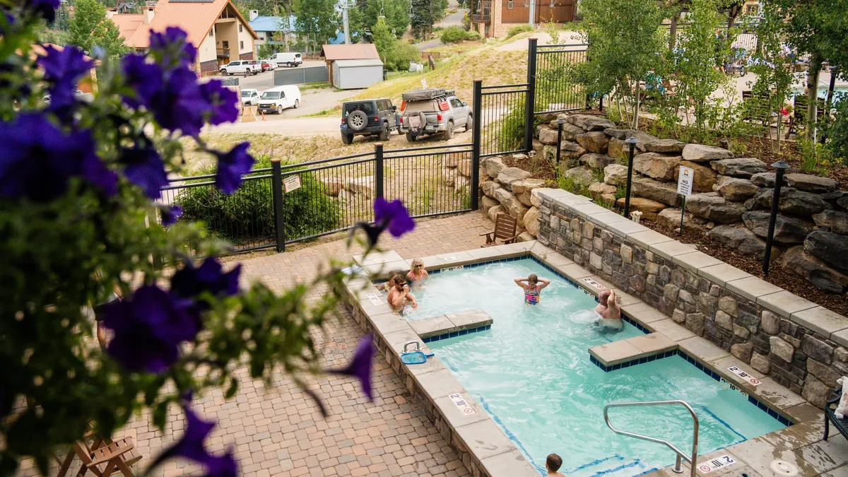 This exterior image captures the resort's swimming pool surrounded by lush flowers, with several guests enjoying the water and relaxing by the poolside. The setting is cheerful and inviting, suggestive of both relaxation and social interaction.