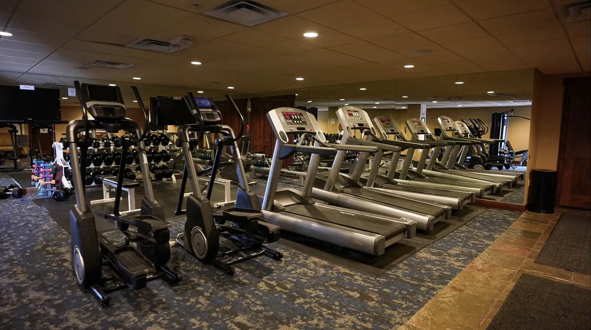 The fitness center is displayed with a row of treadmills, elliptical machines, a rack of dumbbells, and television screens. The atmosphere is modern and motivating, inviting guests to maintain their wellness routines even while on vacation.