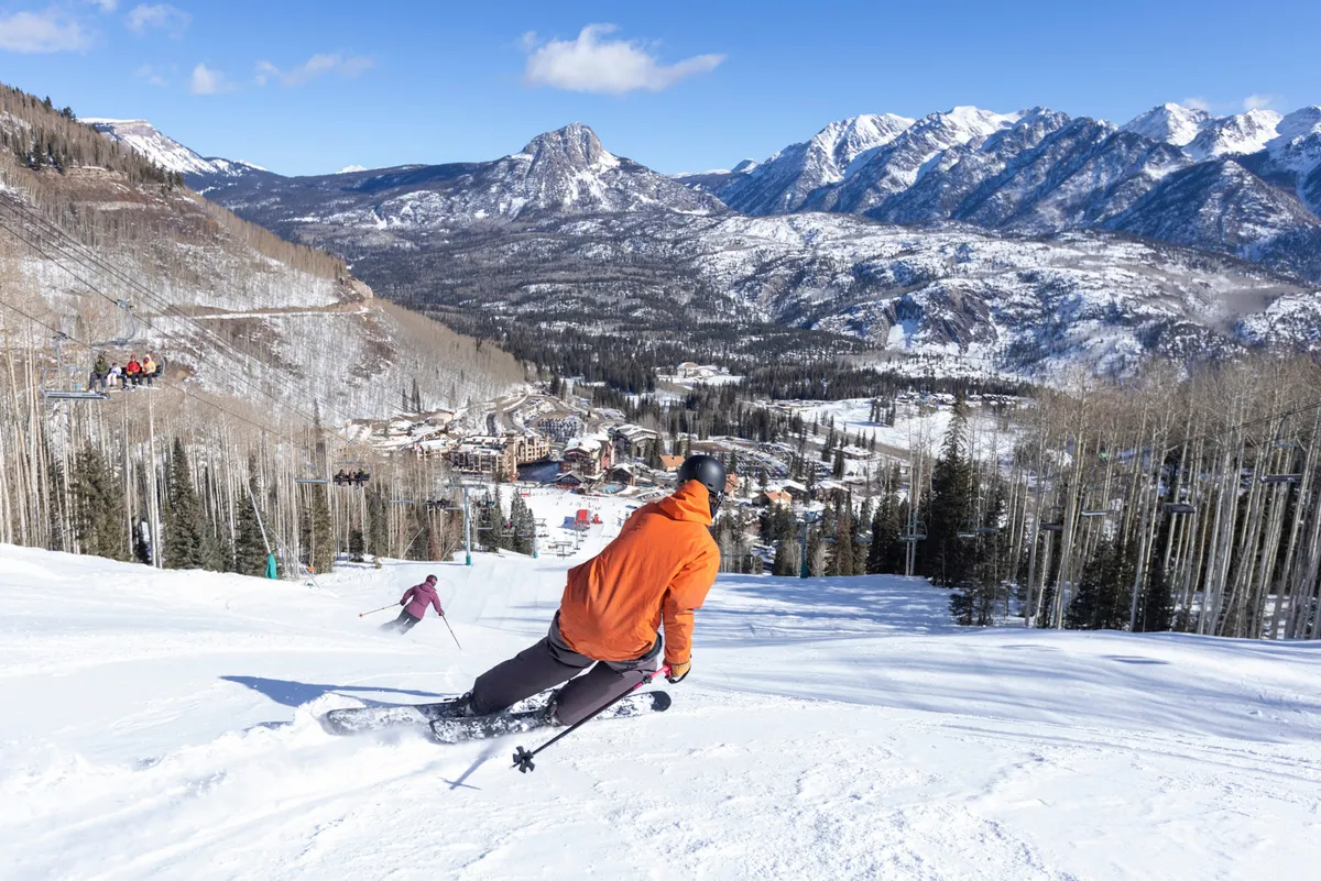 A dynamic perspective from the mountainside, showing two skiers in colorful jackets navigating fresh powder with a panoramic view of the snow-covered Needles Mountain Range and the resort nestled below. The bright blue sky and expansive scenery evoke a sense of freedom and high-altitude adventure.